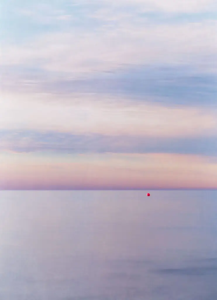Fotografia del silenci, horitzó de mar amb i cel amb colors suaus de posta de sol i una boia vermella surant al mar.